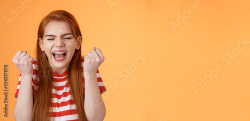 I did it. Charming lucky determined redhead female achieve success, fist pump celebrating, close eyes yell yes yeah, hooray gesture, triumphing victory, very good news, stand orange background photo