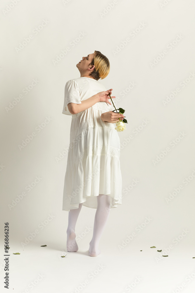 Back view shot of androgynous male model wearing white dress and tights holding white rose behind his back Back view shot of androgynous male model wearing white dress and tights holding white rose behind his back