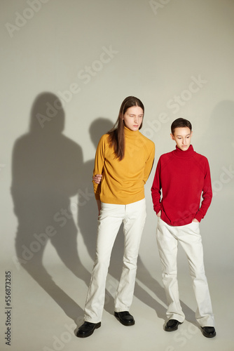 Androgynous man and woman in bright turtleneck sweaters looking away while standing against gray background together