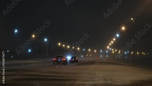 Static shot of cars driving on a well-lit highway during a blizzard with loose snow on the road being blown by strong winds at night photo