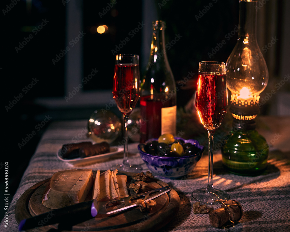 Two glasses with wine, olives in a blue ceramic bowl, cheese with nuts on cheese board on the table in the rays of a kerosene lamp.