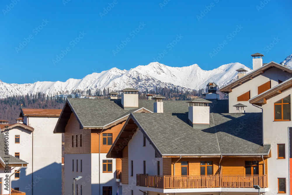 Wooden residential building with sloping roofs in the European style against the backdrop of a mountain snow-capped peak with forests and blue sky.