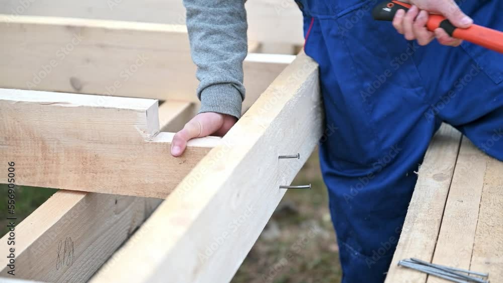 Man worker building wooden frame house on pile foundation. Carpenter hammering nail into wooden board, using hammer. Carpentry concept. Man worker building wooden frame house on pile foundation. Carpenter hammering nail into wooden board, using hammer. Carpentry concept.