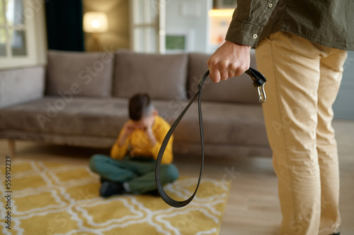 Closeup of father holding leather belt to punish crying son sitting on floor photo