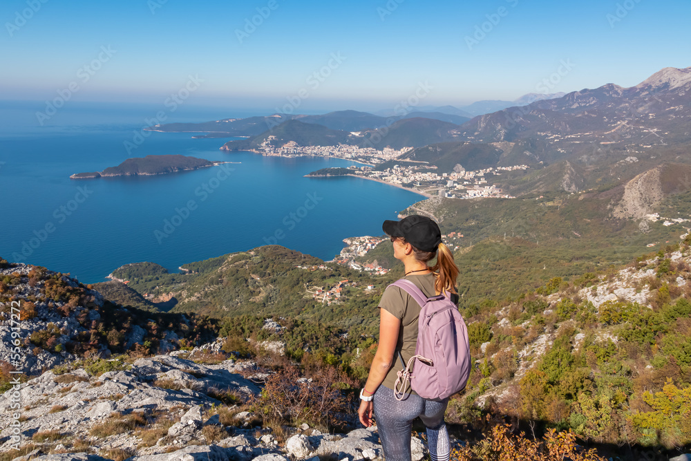 Naklejka premium Hiking woman with backpack enjoying panoramic aerial view on coastline of Budva and Sveti Nikola Island seen from Goli Vrh, Adriatic Mediterranean Sea, Montenegro, Balkan, Europe. Budvanian Riviera