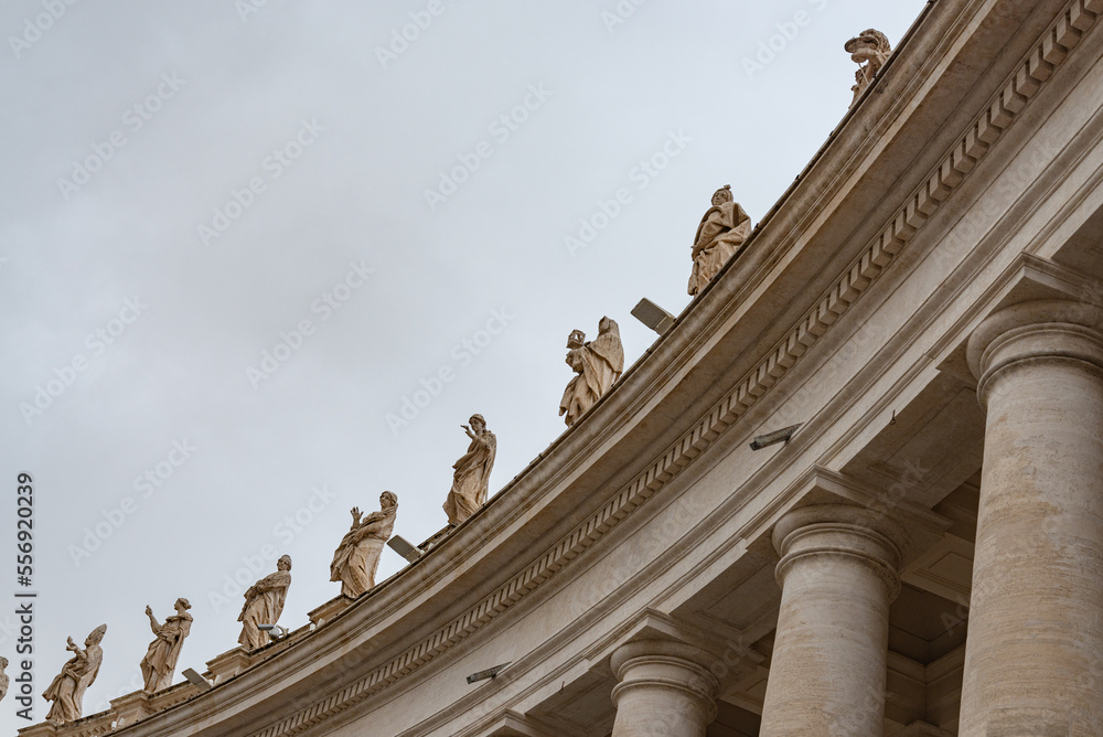 Ancient colonnade with marble statues representing saints in the famous Saint Peter square, in Vatican city, Rome, Italy. Gray sky on the background.