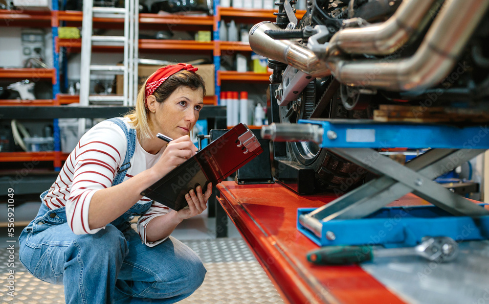 Obraz premium Concentrated mechanic woman with clipboard reviewing damages in motorbike over platform on workshop