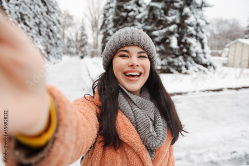 Pretty happy woman in gray hat and pink coat takes selfie and play the ape in winter cold day. Girl takes winter selfie