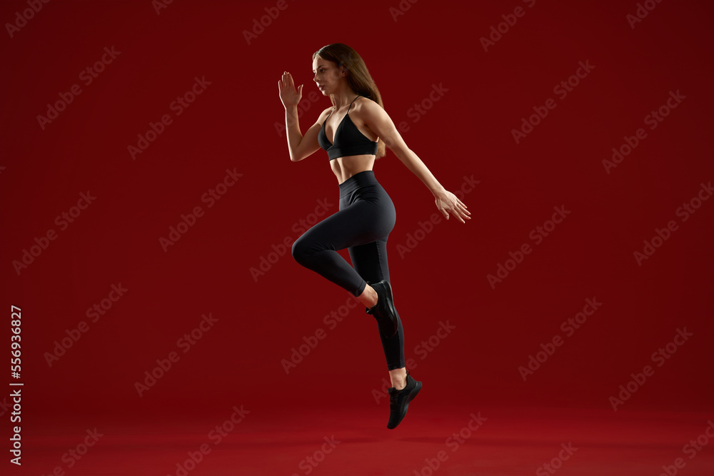 Side view of brunette athletics woman jumping on one leg highly in red studio. Female sports female training showing technique of running or jumping, preparing for competition.