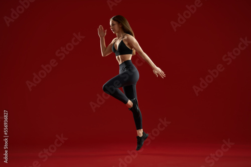 Side view of brunette athletics woman jumping on one leg highly in red studio. Female sports female training showing technique of running or jumping, preparing for competition.