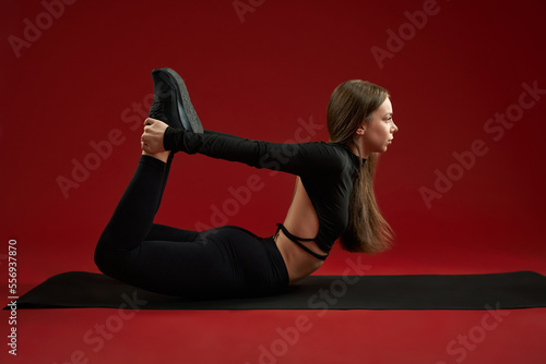 Side view of brunette female doing bow pose on red studio background. Pretty female practice Dhanurasana on black fit mat stretching back and legs. Concept of yoga girl. photo