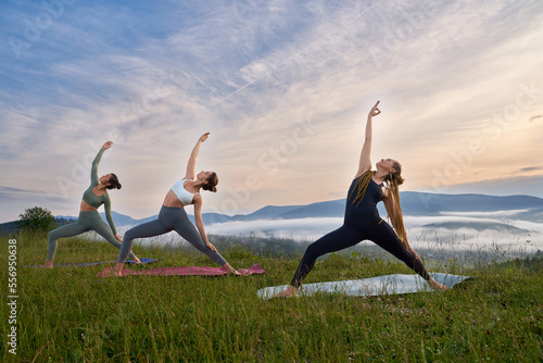 Healthy and sporty three women stretching body on yoga mat during summer day outdoors. Female friends in activewear training together among beautiful mountains.