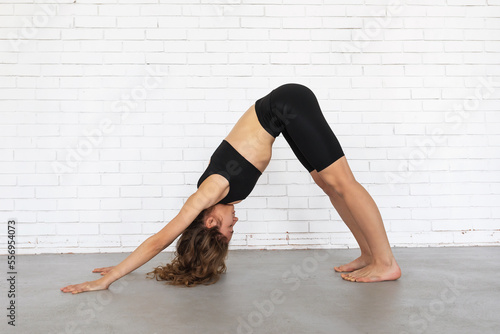 A young woman leading a healthy lifestyle and practicing yoga, performs the exercise of Adho Mukha Schwanasana, a dog pose with her face down, trains in black sportswear near a brick wall photo