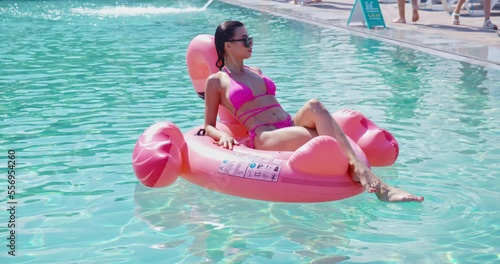 Beautiful young woman using pink inflatable flamingo for swimming in pool. Adorable lady wearing trendy sunglasses and sexy bikini. Relaxation time outdoors. 