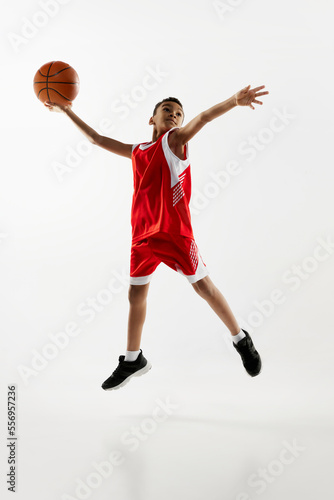 Portrait of boy in red uniform training  playing basketball  throwing ball in a jump over grey studio background. Goal