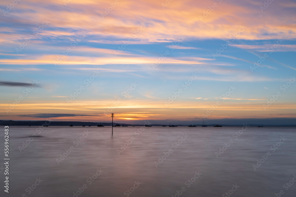Fototapeta premium Summer sunrise over Tenby South beach, Wales, UK. The sun is rising over a calm sea, turning the sky orange and red. There is a single post visible in the ocean.