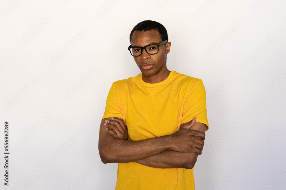 Fototapeta premium Portrait of serious young man wearing glasses posing with folded arms over white background. African American guy wearing yellow T-shirt standing and looking at camera. Confidence concept