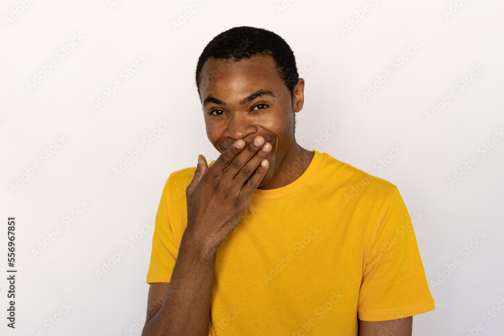 Portrait of positive young man sneering and covering mouth with hand over white background. African American guy wearing yellow T-shirt looking at camera and smiling. Mockery concept Portrait of positive young man sneering and covering mouth with hand over white background. African American guy wearing yellow T-shirt looking at camera and smiling. Mockery concept