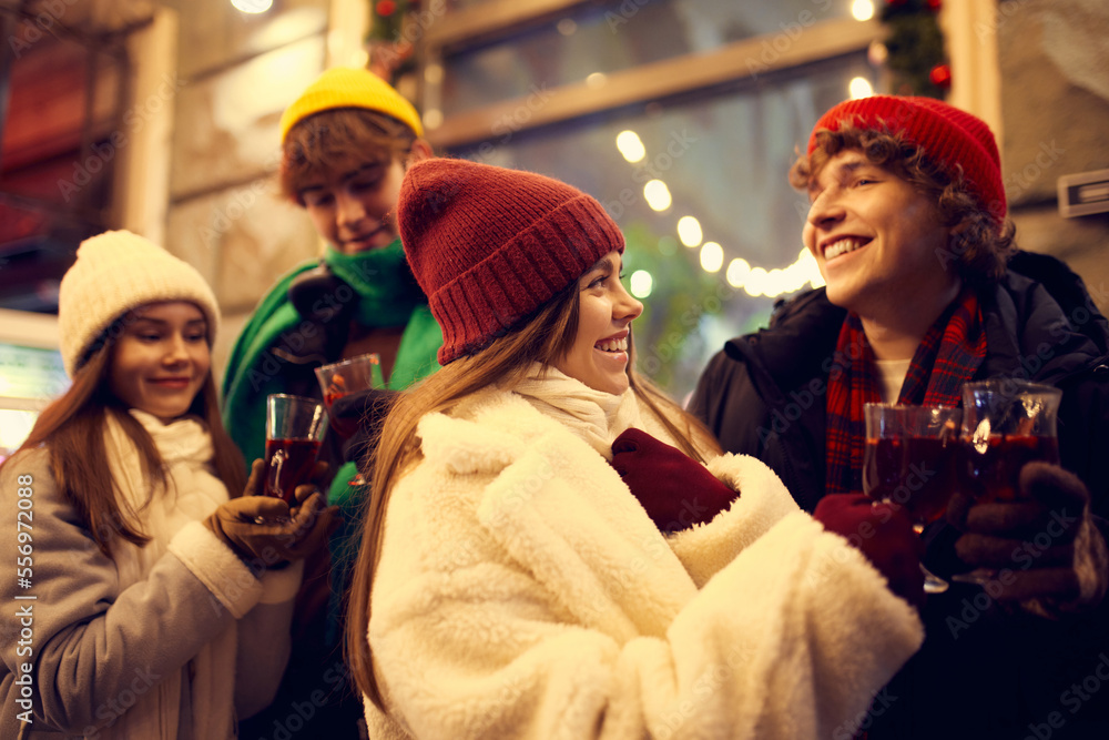 Happy young men and girls, friends walking at festive Christmas street and drinking mulled wine. Concept of holidays, winter, New Years fairs, friendship