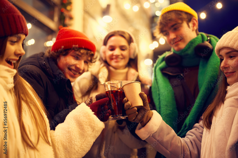 Group of young people, boys and girls spending time together at festive Christmas street, tasting hot mulled wine and coffee. Concept of winter holidays, emotions, friendship