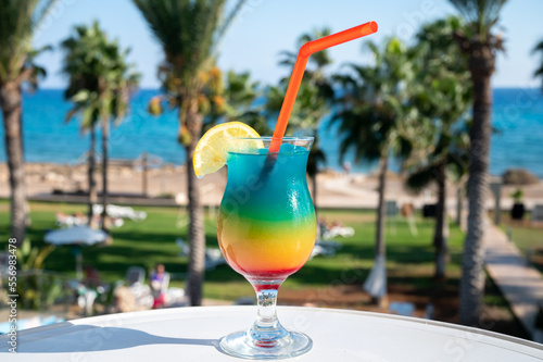 Colourful cold Rainbow Paradise cocktail drink served in glass at pool bar overlooking blue pool, sea and palm trees, relax and holidays at sea