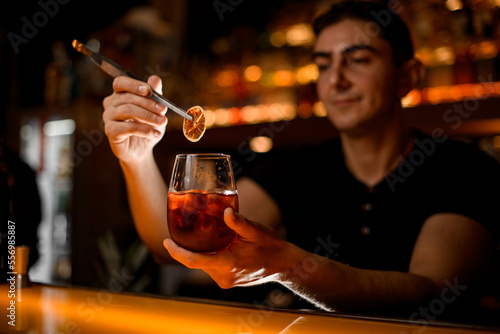 male bartender gently holds slice of dry citrus with tweezers over glass with cold cocktail