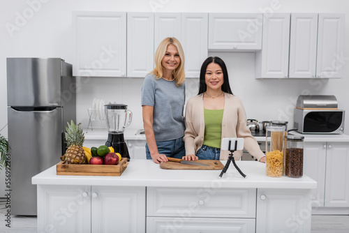 Cheerful interracial bloggers looking at camera near fruits and cellphone on tripod at home