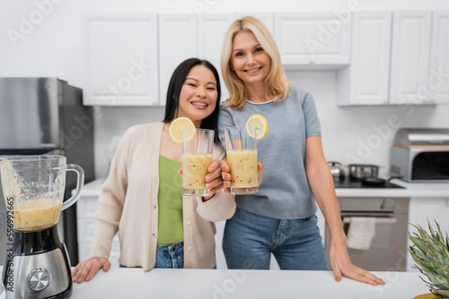 Blurred interracial friends smiling and looking at camera while holding smoothie in kitchen