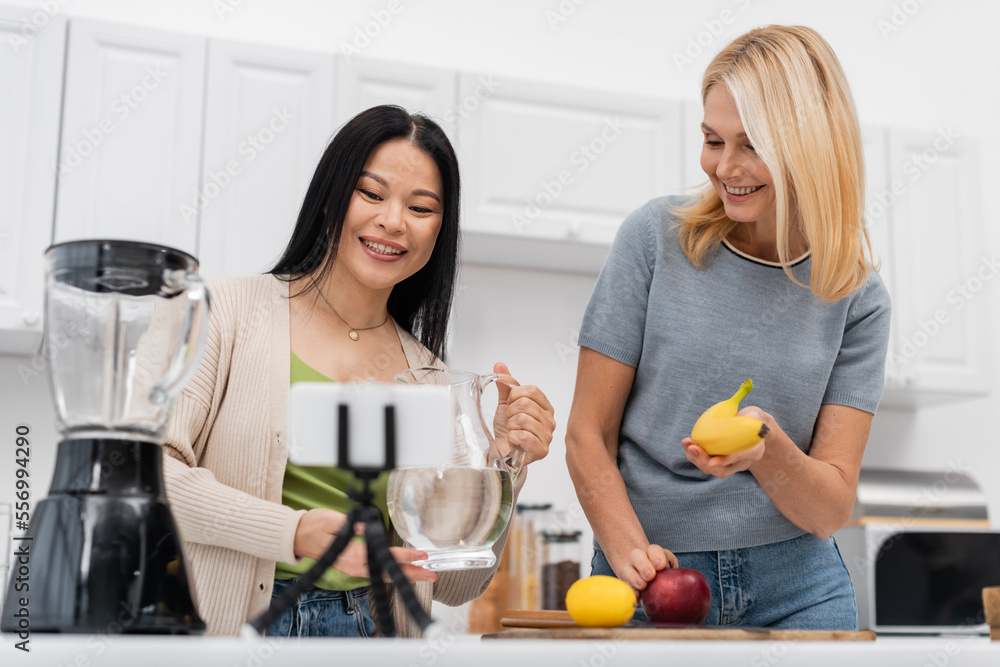 Smiling interracial bloggers holding water and banana near smartphone on tripod at home