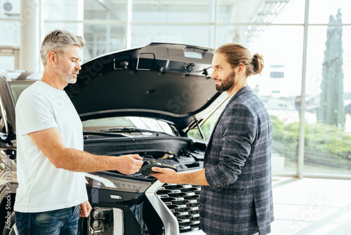 Handsome mature grey haired man buying car from a professional dealer. Successful man buying brand new luxury car in showroom paying via smartphone from mature salesman.