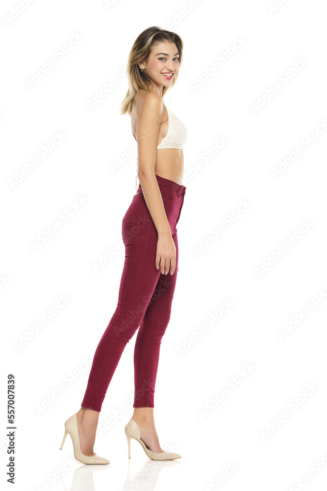 a young smiling woman in burgundy jeans and top, and high heels shoes posing on a white background. Side profile view a young smiling woman in burgundy jeans and top, and high heels shoes posing on a white background. Side profile view