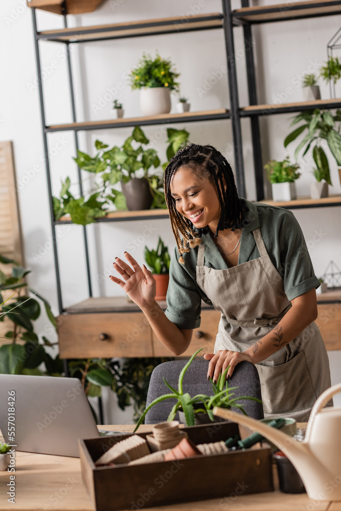 smiling african american florist in apron waving hand near laptop during video call in flower shop smiling african american florist in apron waving hand near laptop during video call in flower shop