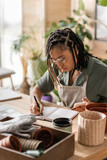 stylish african american florist in apron and eyeglasses writing order in notebook while sitting with smartphone in flower shop