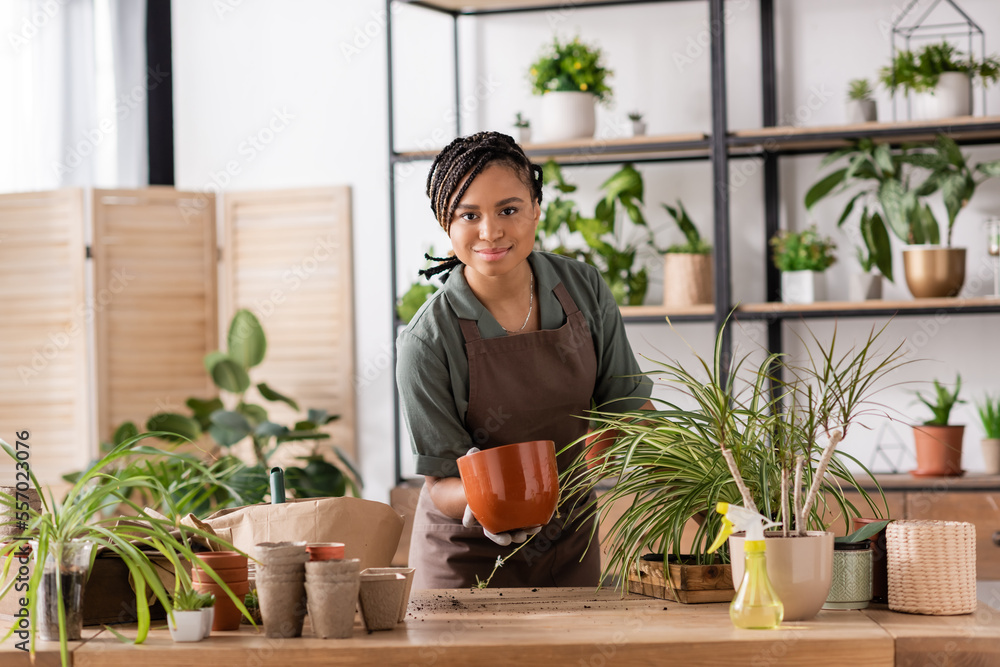 happy african american florist holding flowerpot and looking at camera near plants and spray bottle on table happy african american florist holding flowerpot and looking at camera near plants and spray bottle on table