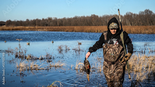 A camouflaged waterfowl hunter retrieves a fallen duck while duck hunting on a sunny day