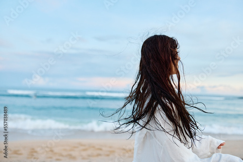 Brunette woman with long hair in a white shirt and denim shorts smile and happiness running on the beach and having fun smile with teeth in front of the ocean, vacation summer trip