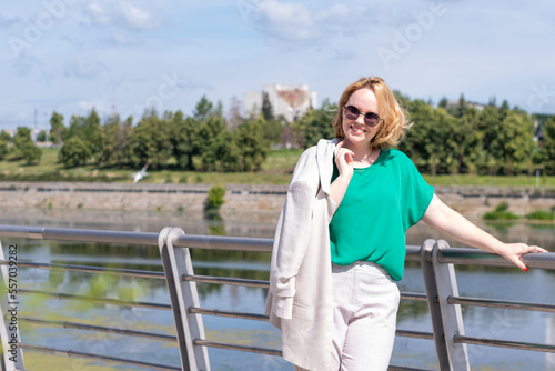 A beautiful pretty girl in a suit holding a jacket in her hand, is standing on the river bank holding on to the railing. © Alena