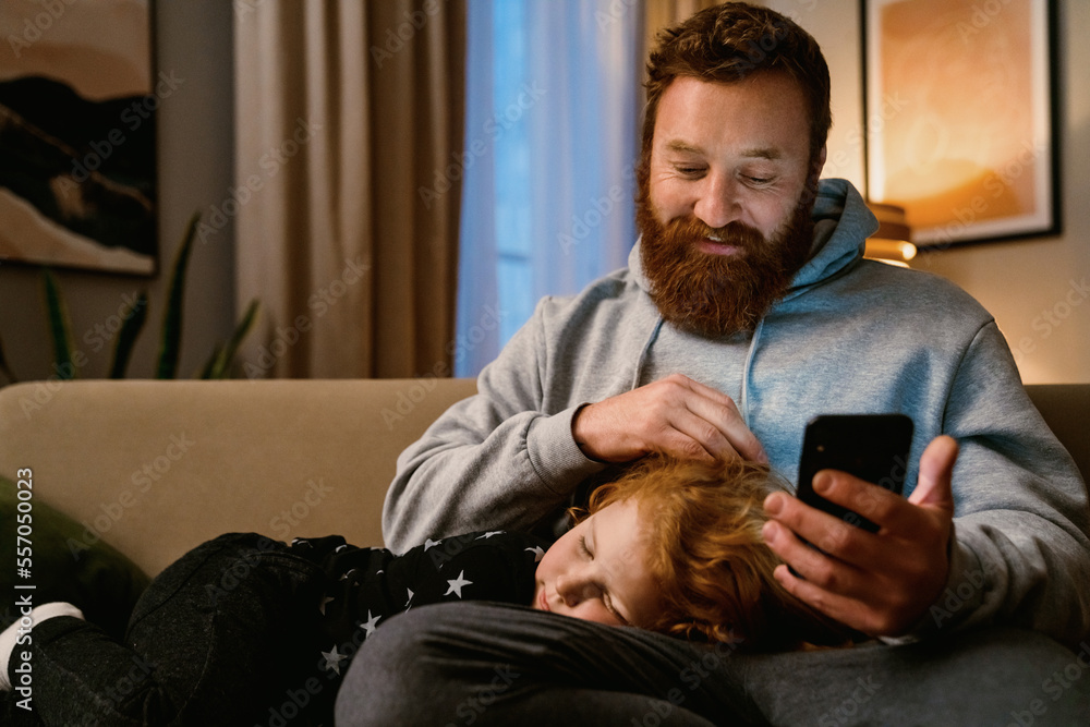 Father using smartphone while his little son sleeping on his knees at home Father using smartphone while his little son sleeping on his knees at home