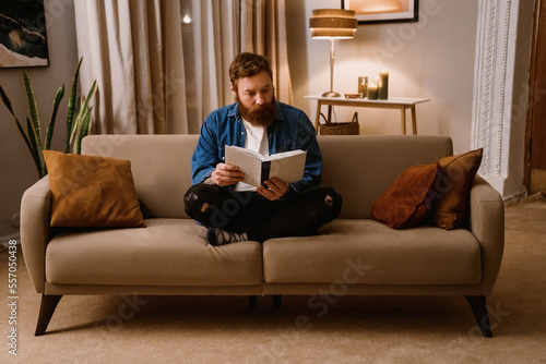 Ginger man reading book while sitting on sofa in living room