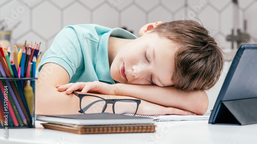 Portrait of young preteen tired boy wearing blue T-shirt, lying sleeping on hands on desk near notebooks, glasses, color pencils, tablet at home. Distant learning, remote education, technology.