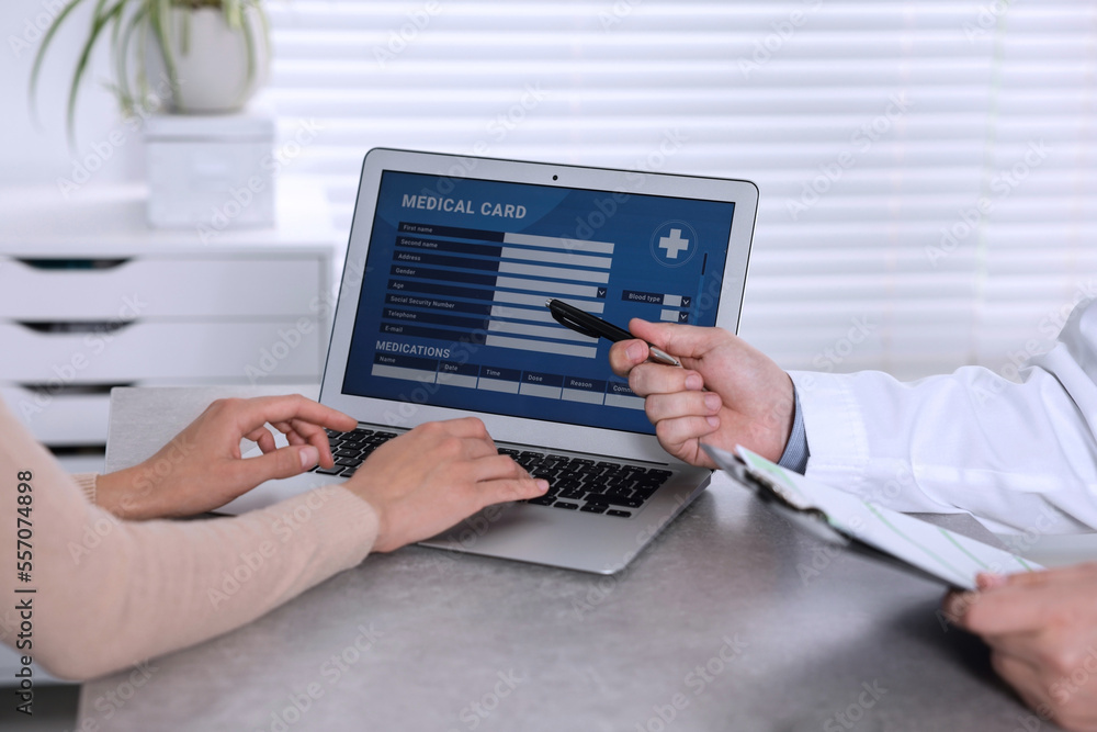 Fototapeta premium Doctor and patient using laptop to fill out medical card at table in clinic, closeup