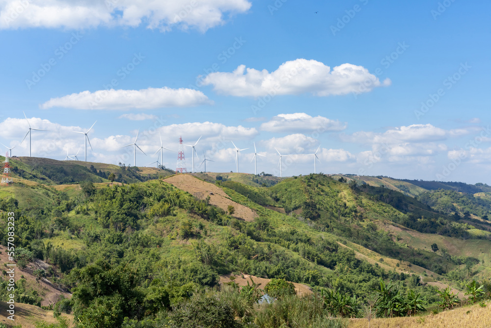 The wind turbines in the top of the grass hill underneath the group of white clouds and blue sky on a sunny day, produced electricity from pure energy.