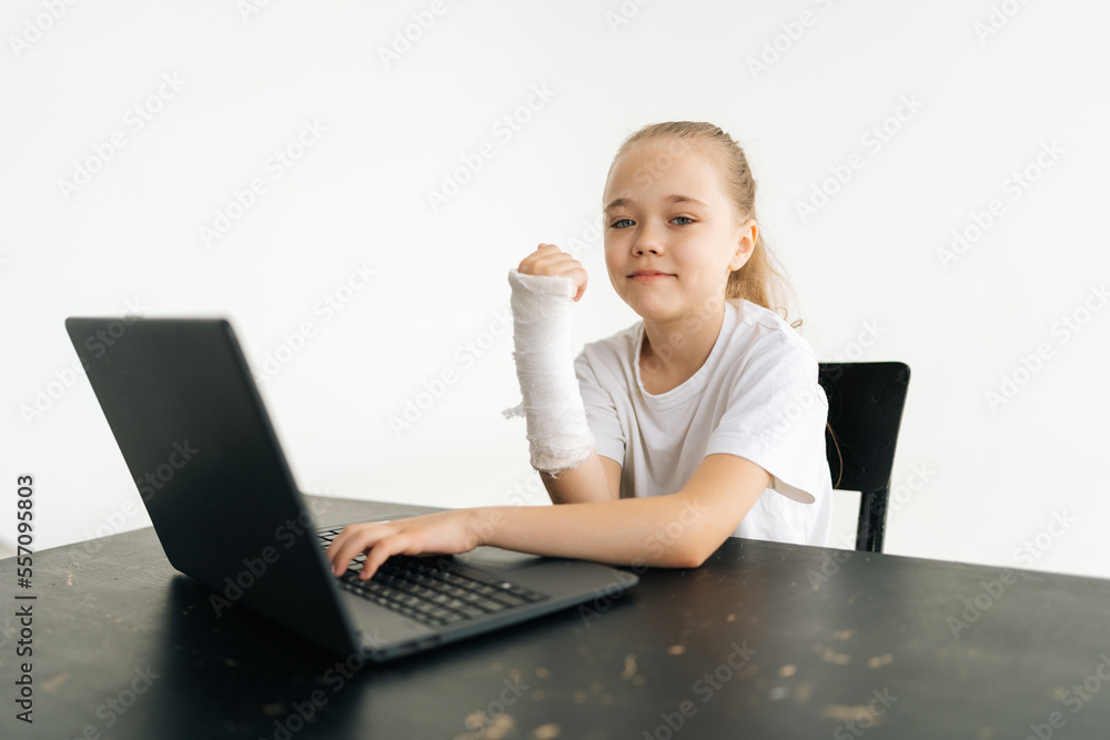 Positive blonde little girl with broken hand wrapped in white plaster bandage sitting at table with laptop computer smiling looking at camera in light room. Concept of child insurance and healthcare. Positive blonde little girl with broken hand wrapped in white plaster bandage sitting at table with laptop computer smiling looking at camera in light room. Concept of child insurance and healthcare.