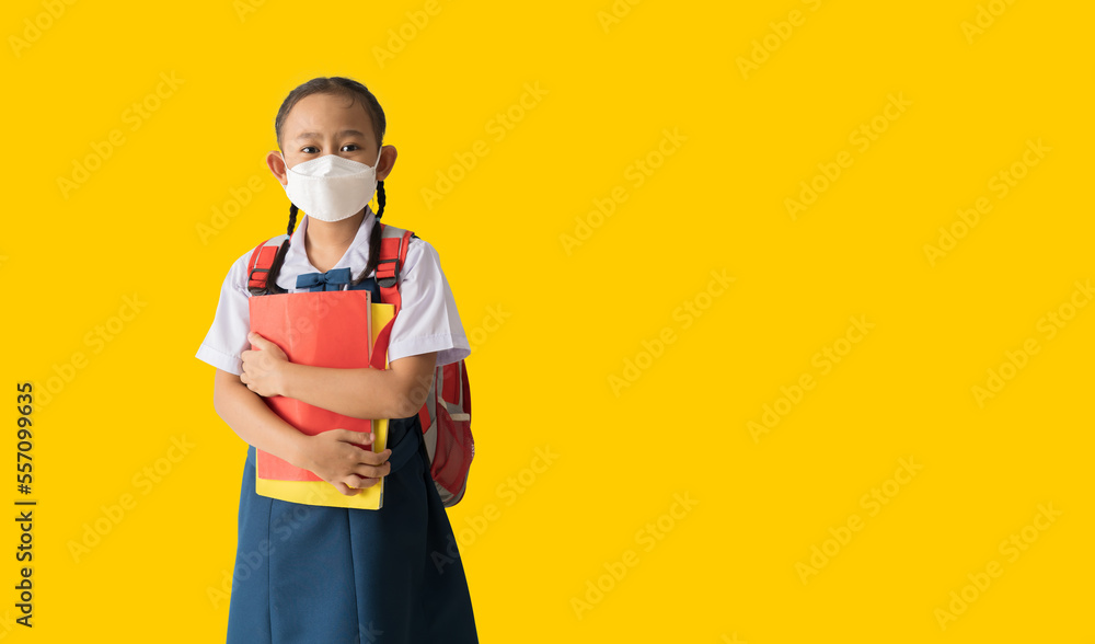 Asian school girl in uniform wear medical face mask, With backpack and holding book isolate on yellow background Asian school girl in uniform wear medical face mask, With backpack and holding book isolate on yellow background