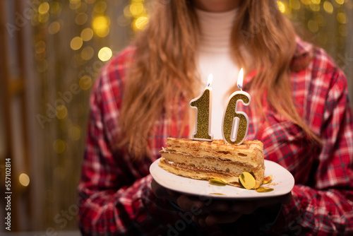 Young woman holding plate with tasty birthday cake with 16 number candle against defocused lights