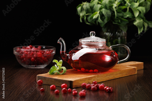 Red herbal tea with cranberries and mint in a glass teapot on a black background. A bunch of mint in the background. Cranberries in the foreground. Selective focus.