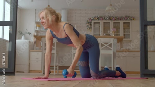 Charming motivated active sporty fit senior woman performing renegade row exercise, toning obliques, rhomboids, and triceps, developing upper body strength while working out at home. photo