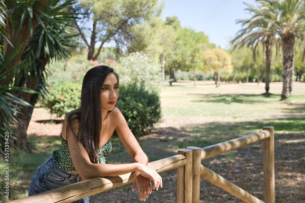 Naklejka premium Young woman, brunette, slender, dressed in green t-shirt and jeans, leaning on a wooden railing, pensive and concentrated. Concept beauty, fashion, trend, thought, concentration.