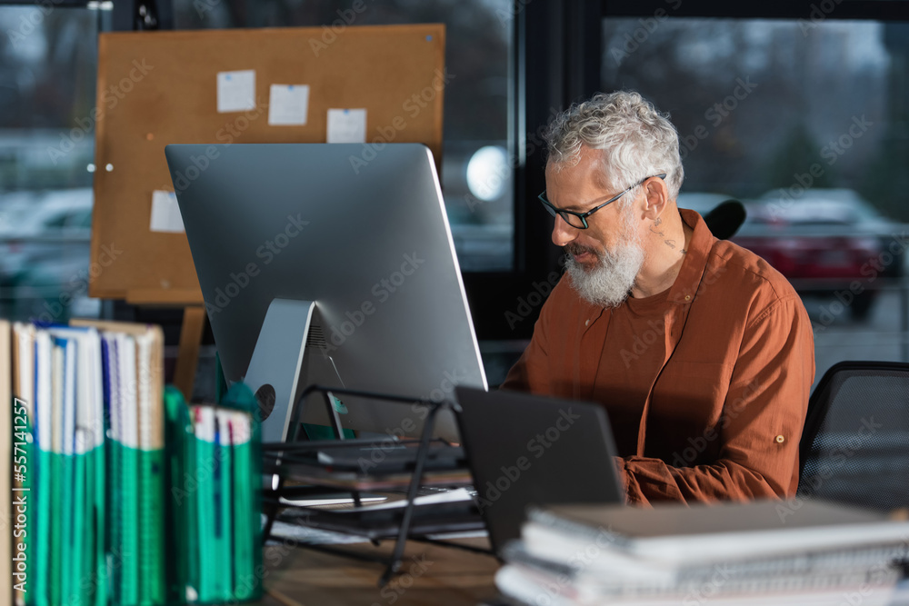 Mature businessman in eyeglasses working near computers and blurred documents in office Mature businessman in eyeglasses working near computers and blurred documents in office