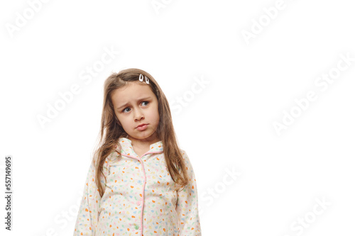 Upset baby girl in pajamas, feeling loneliness while woke up, expressing worried emotion and sadness, looking aside at a copy space for your advertising, standing isolated over white background © Taras Grebinets
